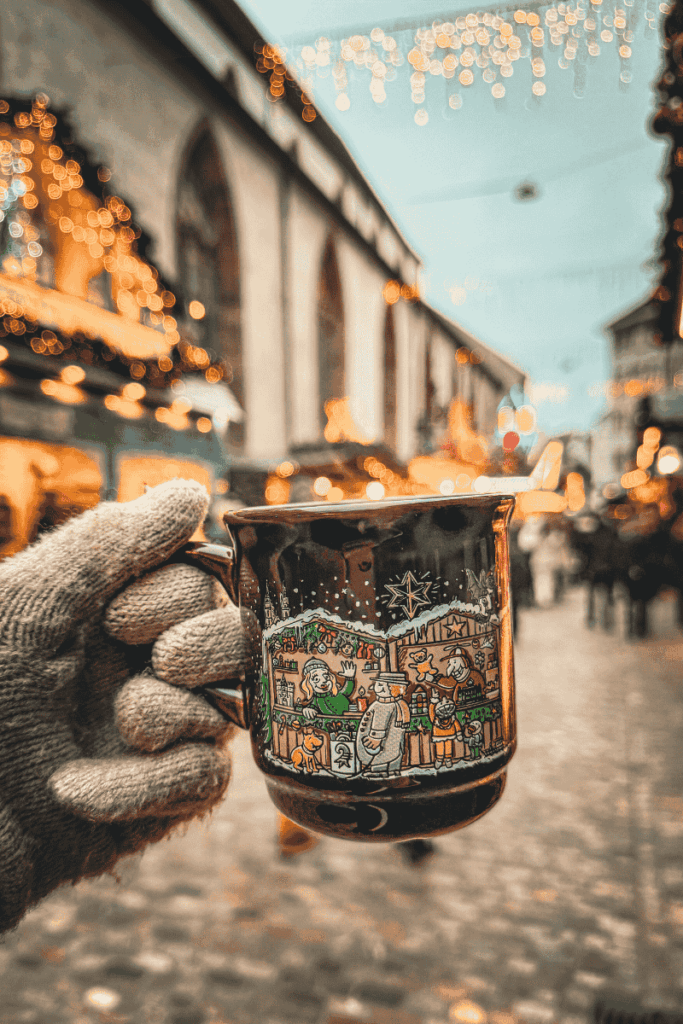 A gloved hand holds a festive ceramic mug decorated with a Christmas market scene, set against twinkling lights and historic buildings on a cobblestone street.