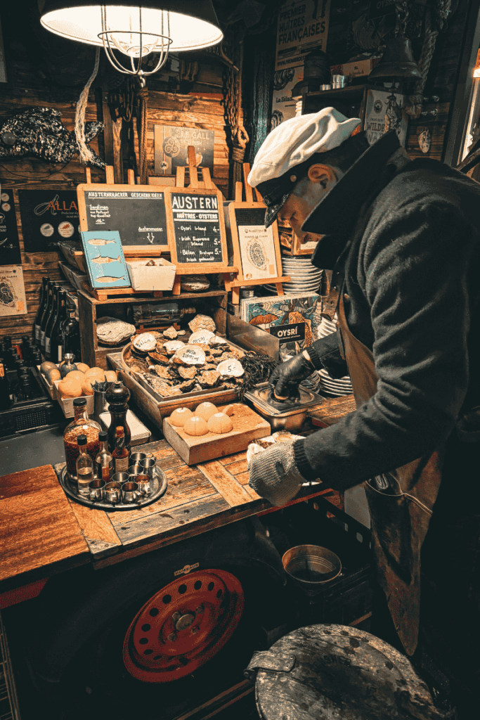 A vendor in a nautical cap prepares fresh oysters at a rustic seafood stall decorated with maritime accents and chalkboard menus at a European Christmas market.