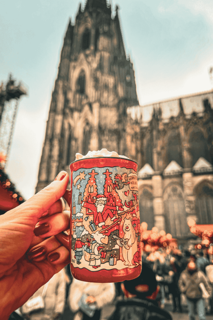 A hand holds a festive red Christmas market mug topped with whipped cream in front of the towering Cologne Cathedral, surrounded by glowing market lights and crowds.
