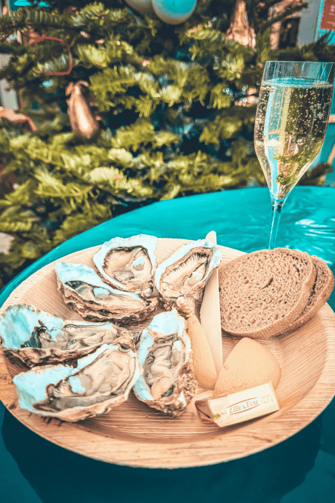 A festive holiday table displays a plate of fresh oysters with lemon wedges, rye bread, and butter, paired with a glass of sparkling wine in front of a decorated Christmas tree.