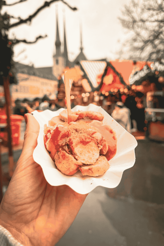A hand holds a paper tray filled with sliced currywurst smothered in ketchup and curry powder, with festive Christmas market stalls and church spires in the background.
