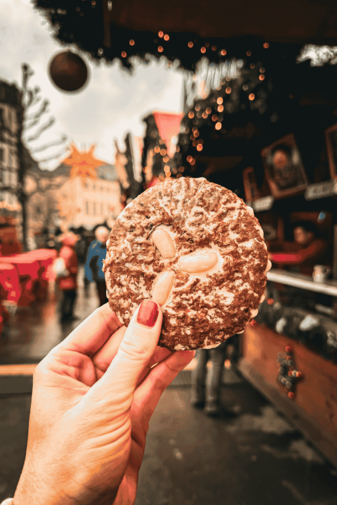 A hand holds up a traditional German Lebkuchen cookie topped with almonds in front of a cozy Christmas market stall decorated with lights and garlands.