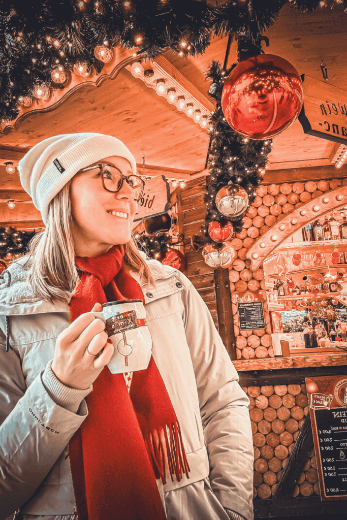 Kate smiles while holding a white mug of mulled wine inside a cozy, festively decorated Christmas market stall adorned with ornaments and warm lights.
