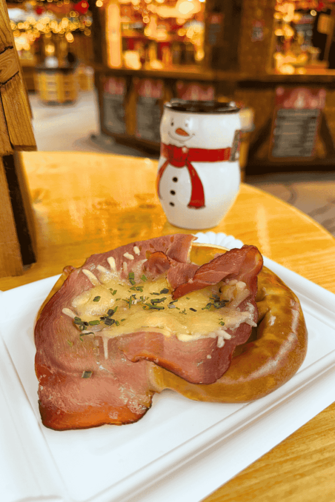 A soft pretzel topped with melted cheese, ham, and chives sits on a white tray beside a festive snowman mug at a cozy Christmas market table.