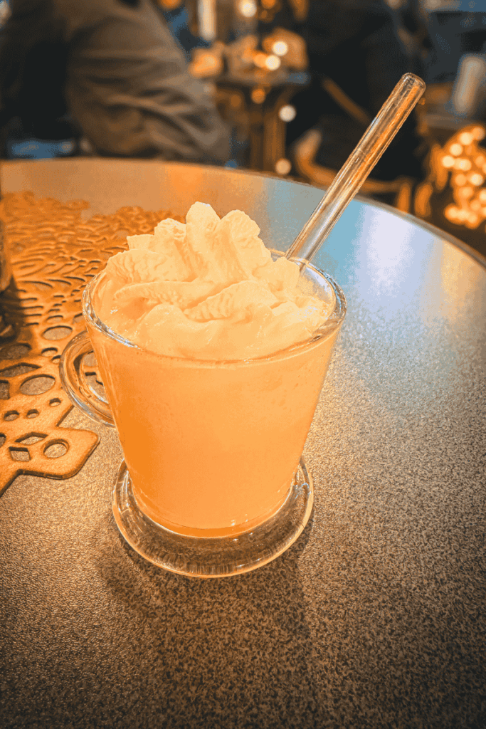 A glass mug filled with creamy Eierpunsch is topped with a generous swirl of whipped cream and served with a clear straw at a cozy Christmas market café table.