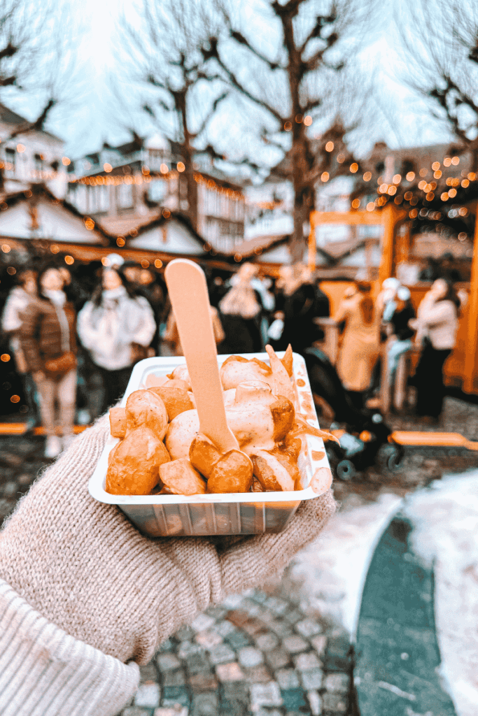 A gloved hand holds a tray of Champignons (mushrooms) smothered in creamy garlic sauce, with a festive Christmas market crowd in the background.