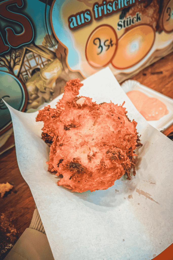A freshly fried potato pancake, golden and crispy, rests on parchment paper with a side of pink dipping sauce at a Christmas market food stall.