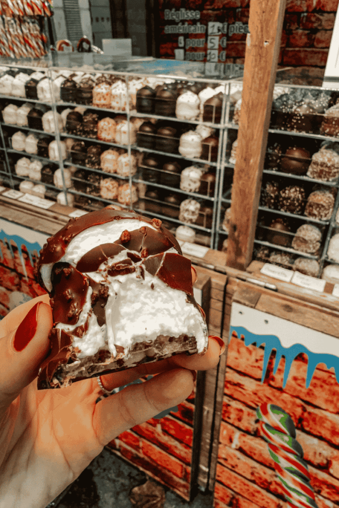 A hand holds a bitten Schokokuss—fluffy marshmallow filling inside a crisp chocolate shell—in front of a display case filled with assorted chocolate-covered treats at a Christmas market.