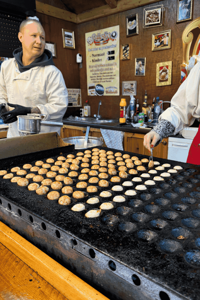Vendors prepare rows of golden poffertjes—mini Dutch pancakes—on a large griddle at a bustling Christmas market stall.