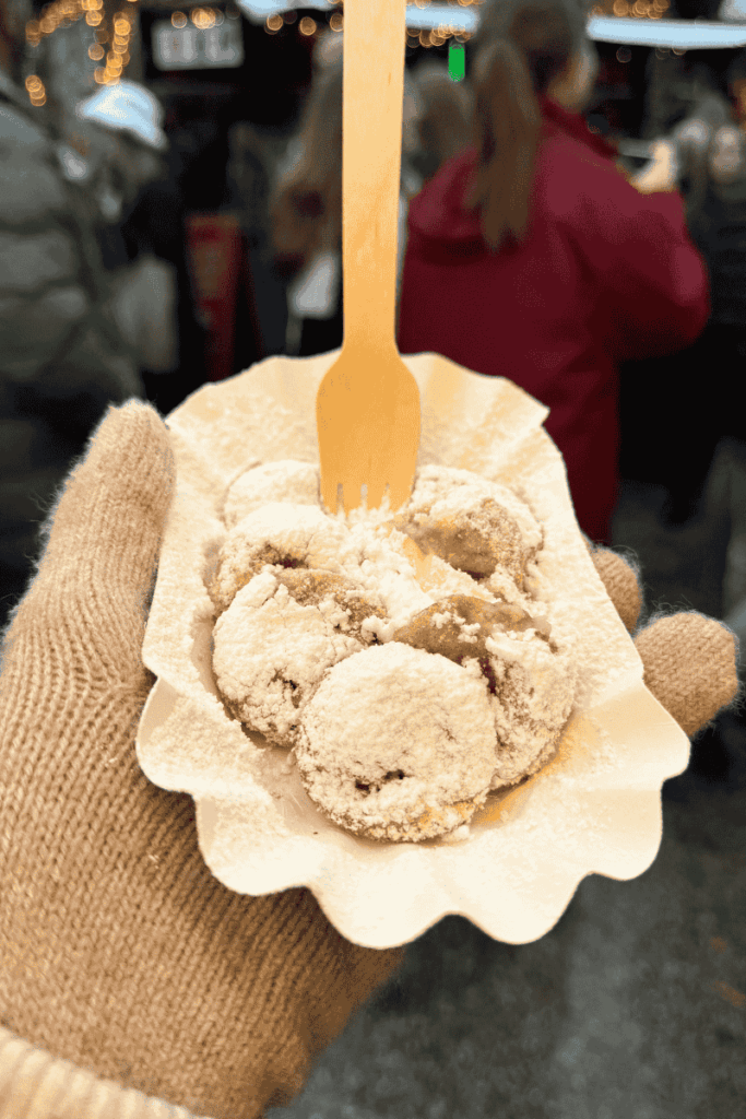 A gloved hand holds a tray of warm poffertjes dusted with powdered sugar and served with a wooden fork at a festive holiday market.