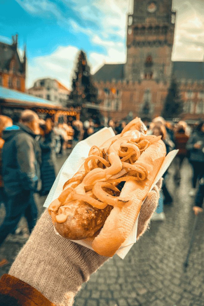 A gloved hand holds a hearty bratwurst in a bun topped with grilled onions, with a Christmas market and historic clock tower in the background.