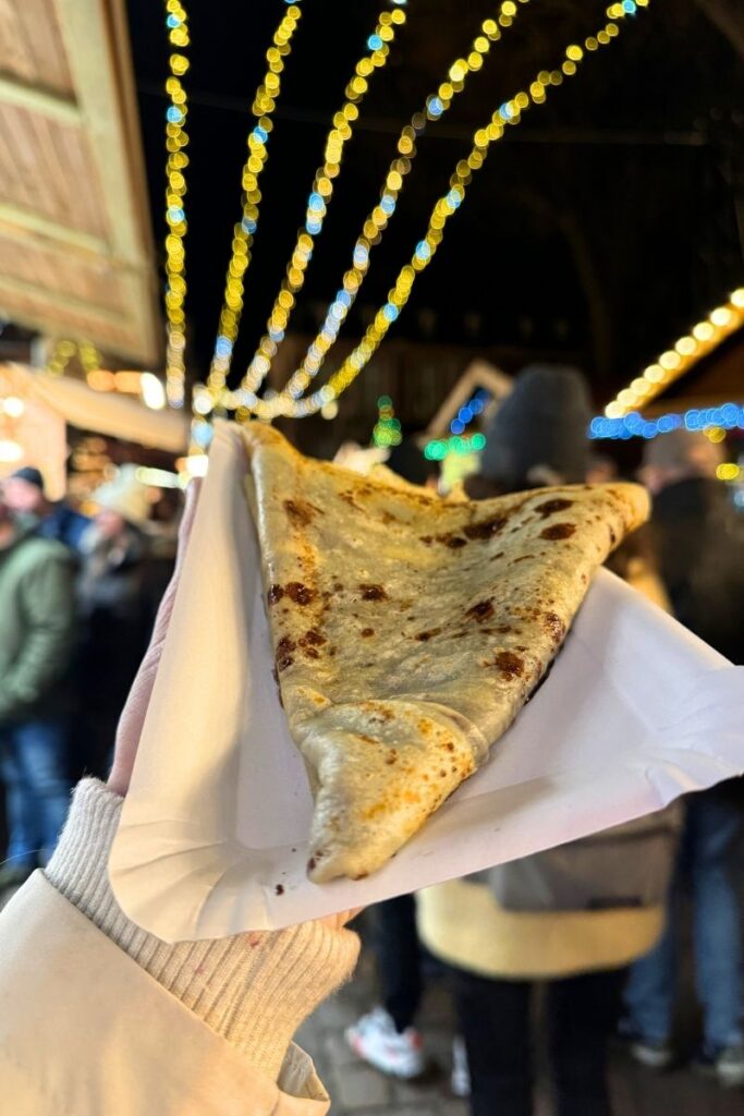 A hand holds a freshly made folded crepe in a paper tray beneath festive string lights at a bustling Christmas market at night.
