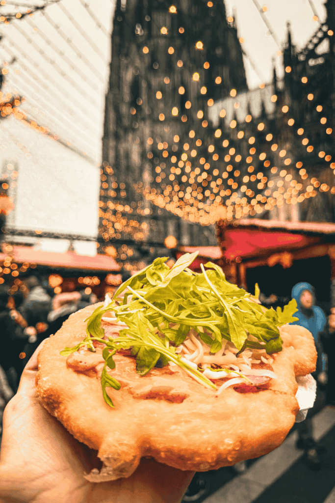 A hand holds up Hungarian lángos topped with arugula, bacon, and cheese beneath twinkling string lights at a bustling Christmas market near a towering Gothic cathedral.