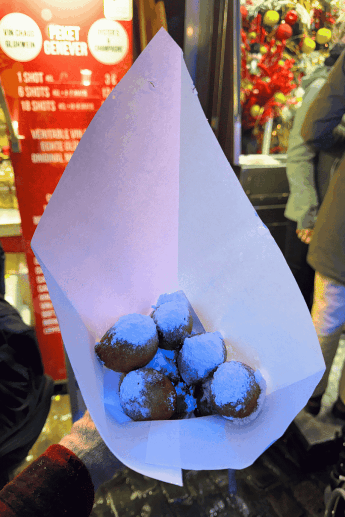 A paper cone holds a serving of warm, golden Mutzenmandeln generously dusted with powdered sugar, glowing under colorful Christmas market lights.