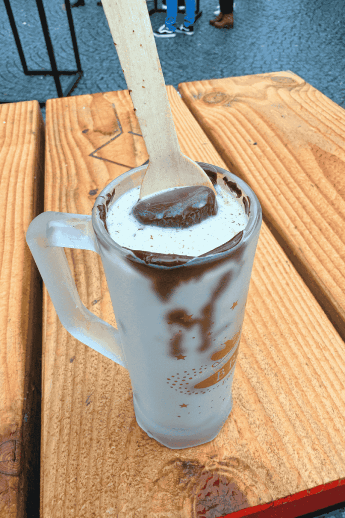 A clear mug filled with rich white hot chocolate is topped with a thick swirl of melted dark chocolate and served with a wooden spoon at a Christmas market table.