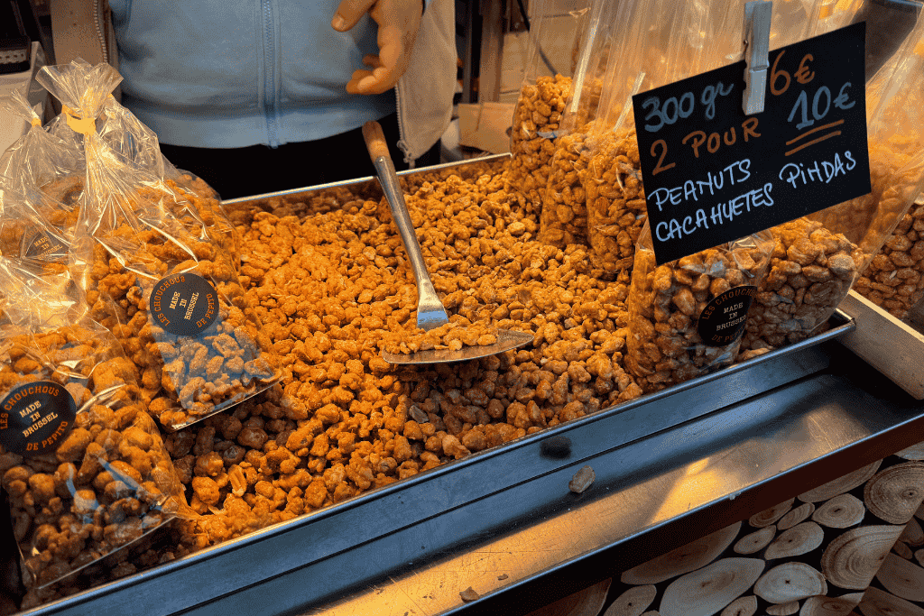 A festive market stall displays a large tray filled with caramelized peanuts, with pre-packed bags labeled “Les Chouchous” and a handwritten sign listing prices in multiple languages.
