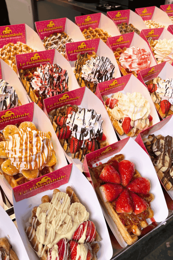 An eye-catching display of Belgian waffles from Le Funambule, each topped with combinations of whipped cream, strawberries, bananas, chocolate drizzle, and caramel sauce in festive pink packaging.