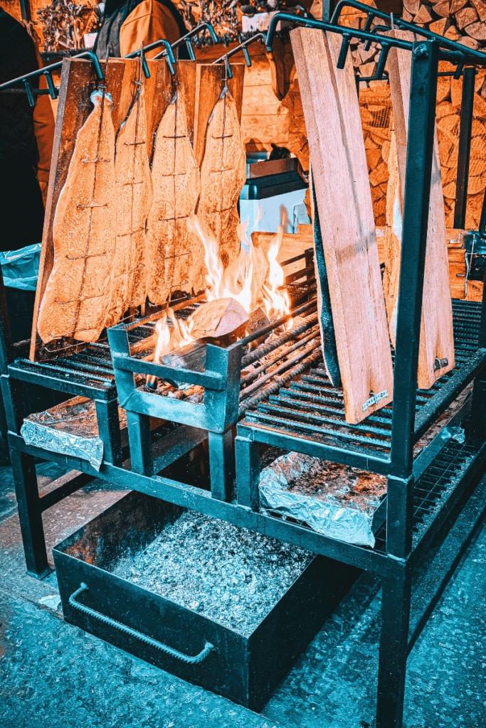 Salmon fillets nailed to wooden planks are slow-cooked vertically over an open fire in a rustic smoker setup at a festive food stall.