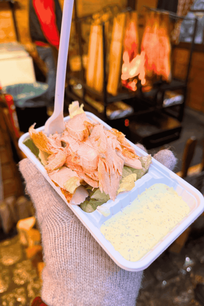 A tray of freshly flaked hot smoked salmon served over lettuce with a side of creamy honey mustard, held by a gloved hand at a Christmas market.
