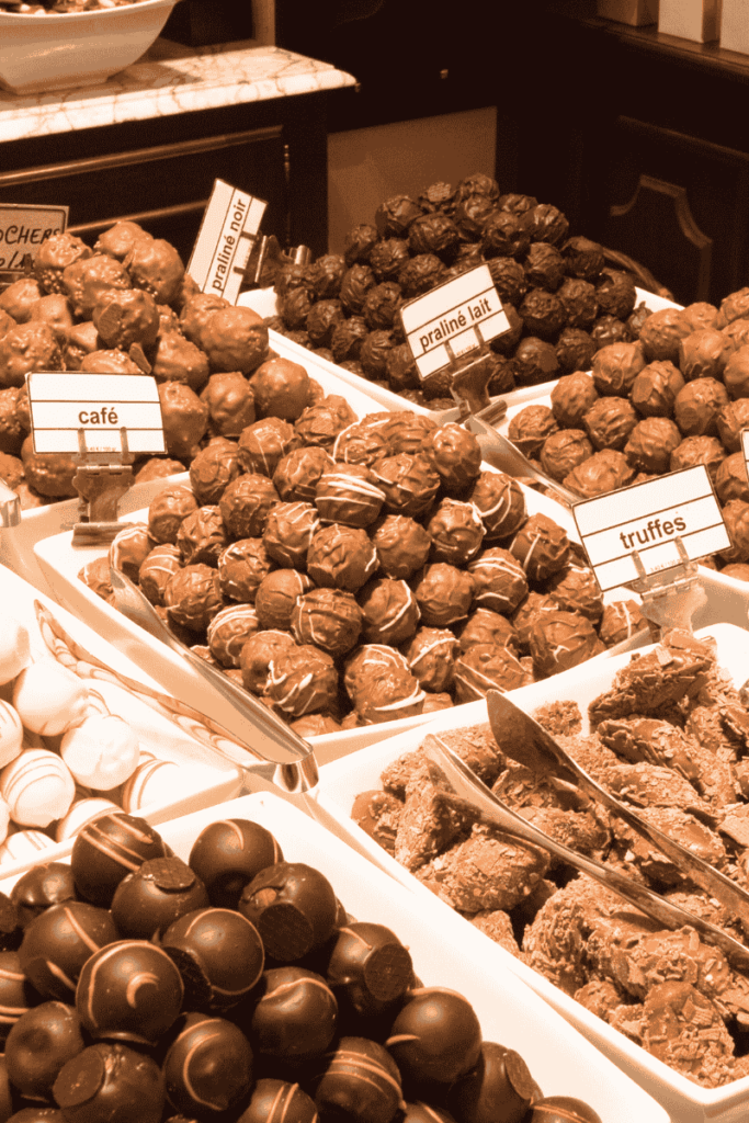 A decadent display of assorted Belgian chocolates and truffles labeled with flavors like café, praline lait, and truffes, arranged in white trays with metal tongs for serving.