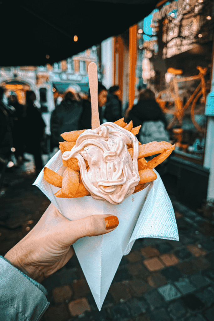 A cone of crispy Belgian fries topped with a swirl of creamy sauce, held by Kate against the bustling backdrop of a Christmas market.