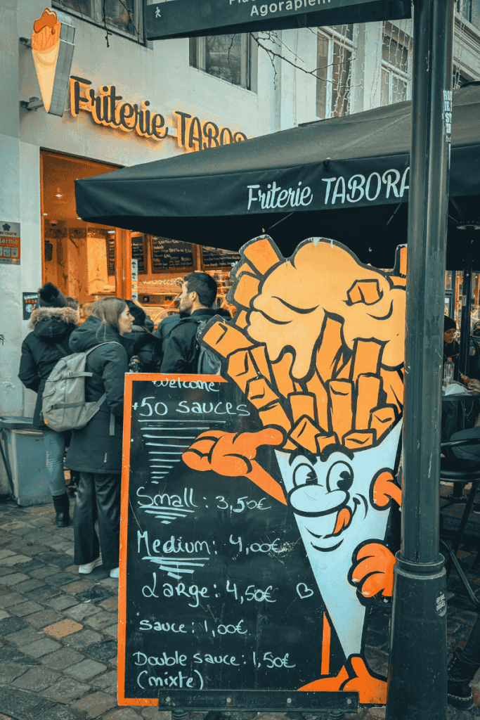 The entrance to Friterie Tabora features a cheerful cartoon cone of fries and a chalkboard menu boasting over 50 sauces and price options.