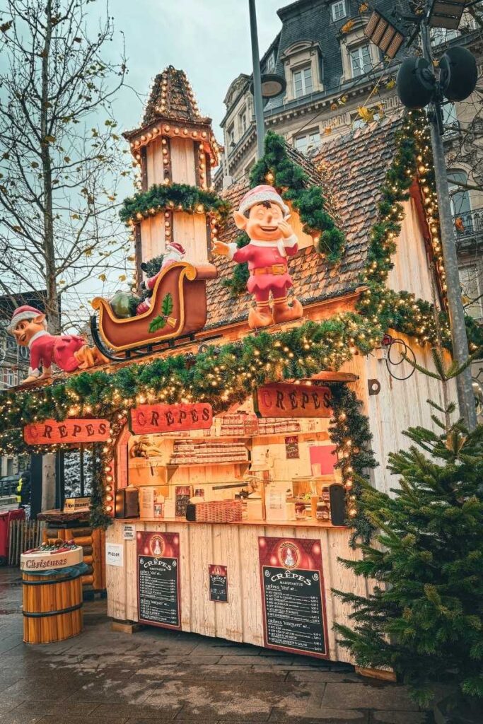 A whimsical Christmas market crepe stall is decorated with garlands, twinkling lights, and playful elf figures, with a sleigh perched on the roof.