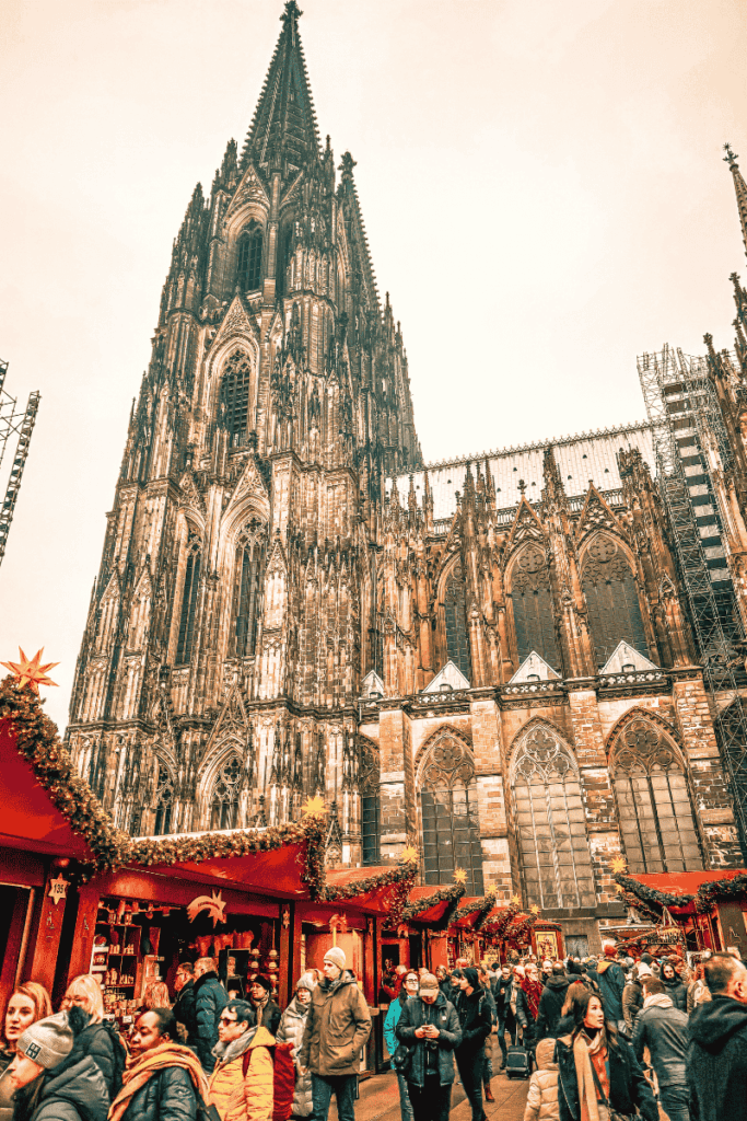 Crowds explore festive Christmas market stalls at the base of the towering Cologne Cathedral, surrounded by garlands and Gothic architecture.