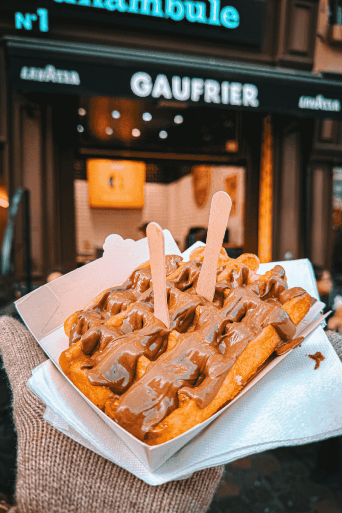A warm Belgian waffle generously drizzled with melted milk chocolate is held in front of the Gaufrier storefront at Le Funambule in Brussels.