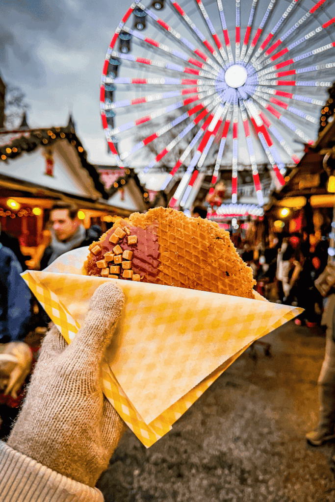 A hand holds a stroopwafel dipped in chocolate and sprinkled with caramel cubes against the glowing backdrop of a Christmas market ferris wheel.