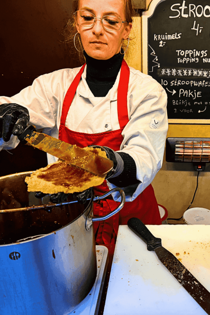 A vendor in a red apron spreads warm caramel filling onto a freshly baked stroopwafel at a market stall.
