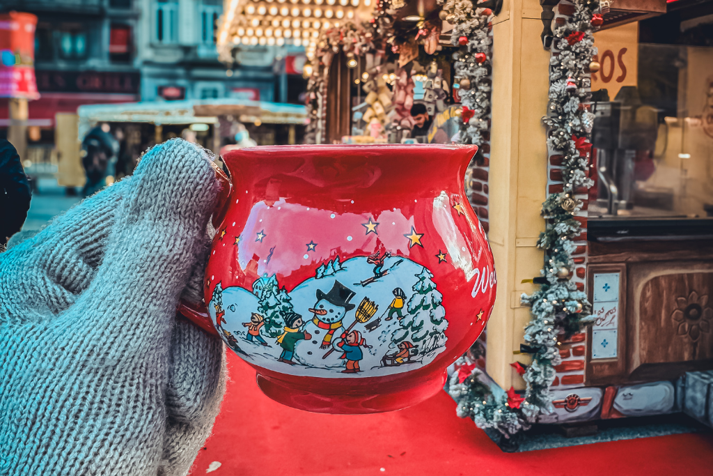 A gloved hand holds a festive red mug decorated with snowmen and Christmas trees, filled with steaming glühwein at a holiday market stall.