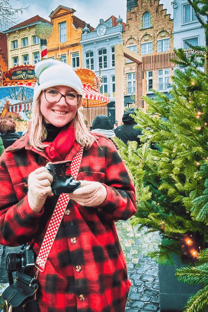 Kate smiles while holding a festive boot-shaped mug of glühwein at a European Christmas market, surrounded by decorated evergreens and colorful historic buildings.