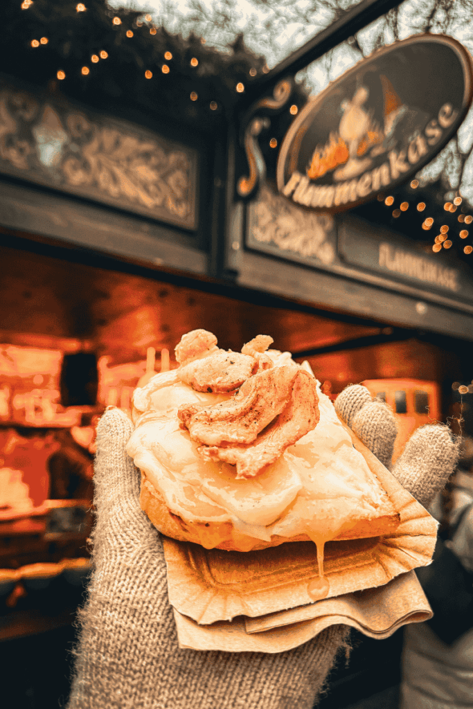 A gloved hand holds up a slice of Flammekäse bread smothered in melted cheese and crispy bacon in front of a festive Christmas market stall.