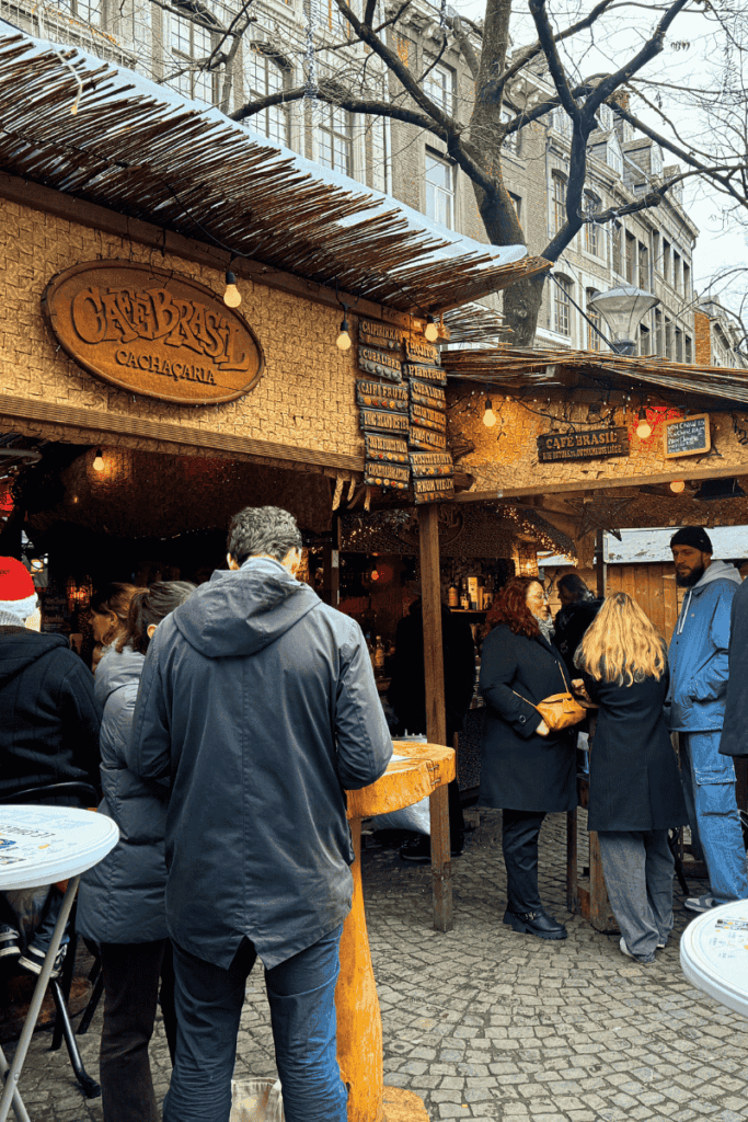 People gather around the rustic Café Brasil stall at a Christmas market, which offers Brazilian cocktails like caipirinhas and cachaça-based drinks under a canopy of string lights.