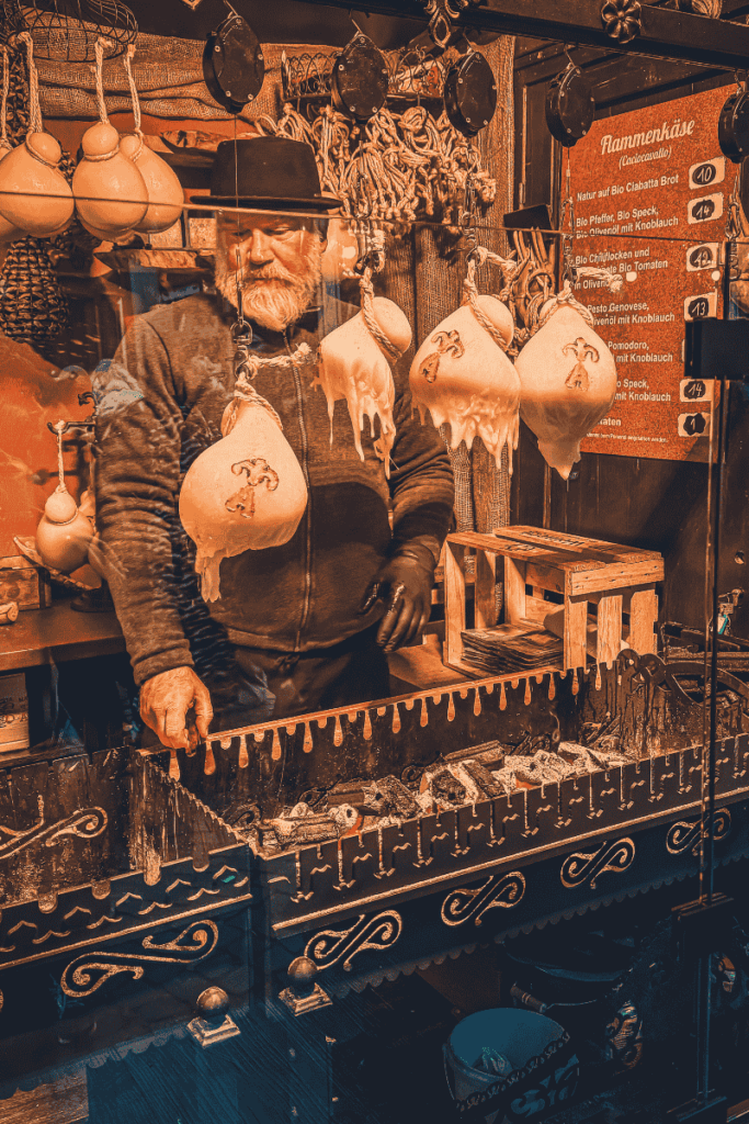 A vendor in traditional attire tends to a rustic stall selling Flammekäse, surrounded by hanging cheese rounds and a wood-fired grill.