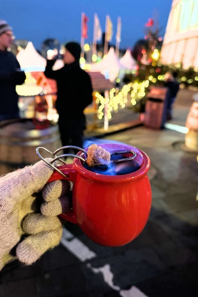 A gloved hand holds a red mug of flaming Feuerzangenbowle, with a sugar cube set ablaze above the spiced mulled wine at a festive Christmas market at night.