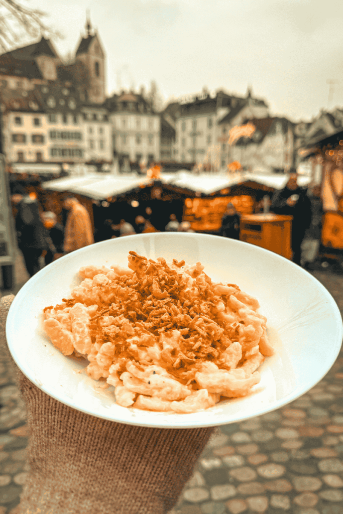 A hand holds a white bowl of creamy käsespätzle topped with crispy fried onions above a charming Christmas market scene in a historic European town.