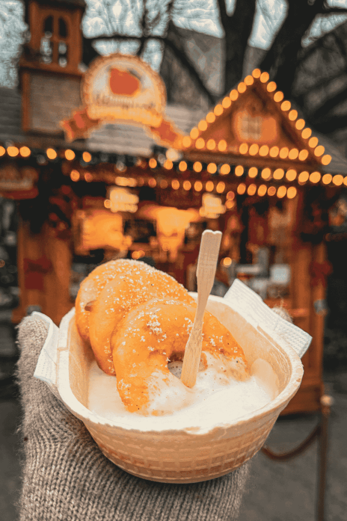 A gloved hand holds a tray of golden fried apple rings dusted with powdered sugar and served with vanilla sauce in front of a warmly lit Christmas market stall.