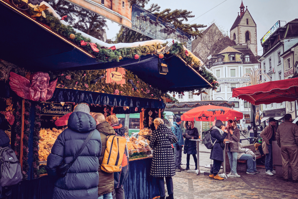 Crowds gather at a festive Christmas market stall selling assorted pastries and holiday sweets, with visitors enjoying food under a red umbrella.