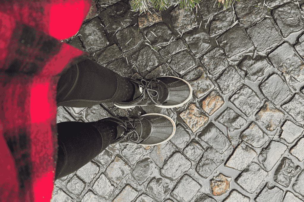 Kate looks down at her waterproof winter boots standing on glistening cobblestone streets, wet from rain and bordered by evergreen branches.