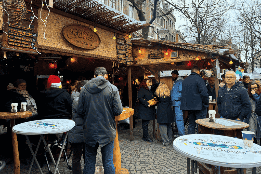 Crowds gather around a cozy wooden stall labeled “Café Brasil Cachaçaria” at a European Christmas market, sipping warm drinks beside round standing tables under festive string lights.