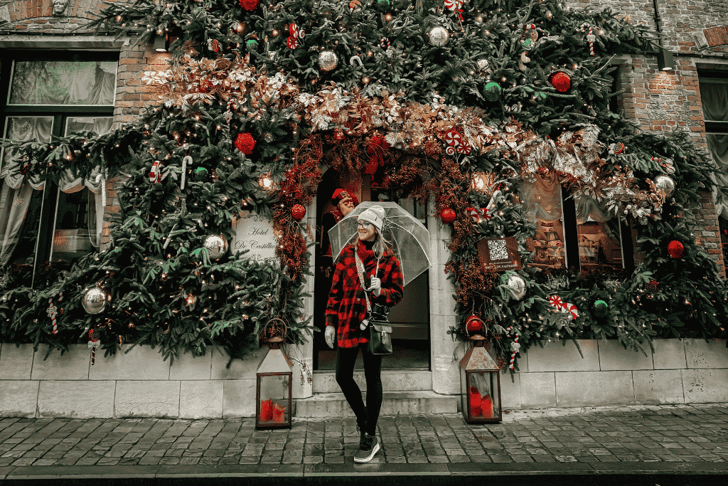 Kate stands under a clear umbrella in front of a hotel entrance lavishly decorated with Christmas garlands, ornaments, and greenery spilling across the brick façade.