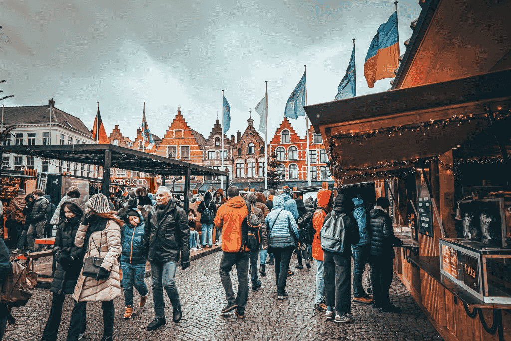 A bustling Christmas market square filled with bundled-up visitors and food stalls, set against a row of Dutch-style buildings topped with colorful flags.