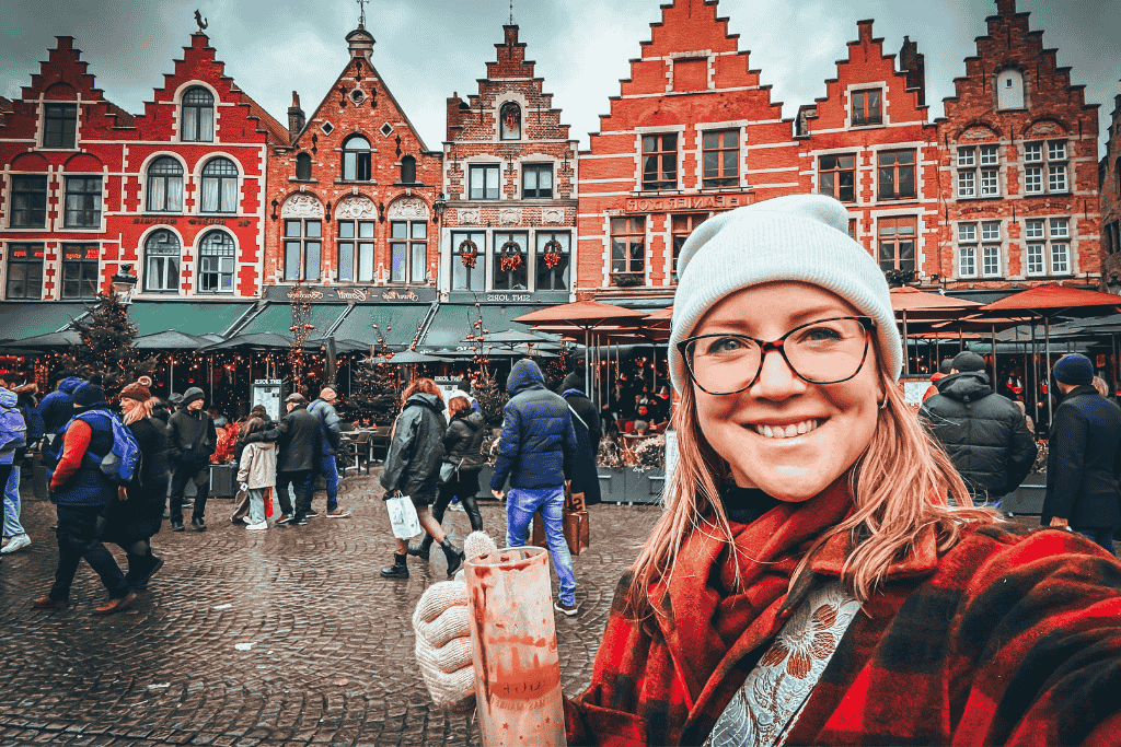 Kate smiles while holding a festive drink in the bustling Bruges market square, surrounded by colorful stepped-gable buildings and holiday crowds.