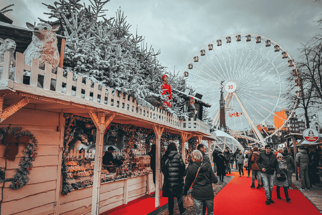 Visitors, including Kate, stroll past snow-dusted market stalls and festive decorations under a towering Ferris wheel at a bustling European Christmas market.