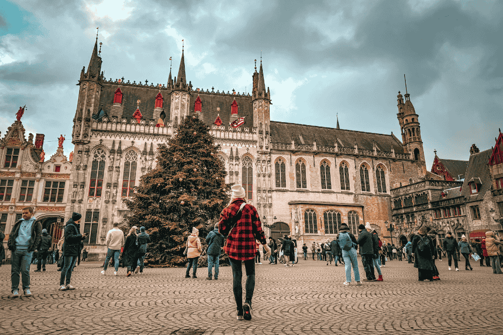 Kate walks toward a large Christmas tree in a historic European square surrounded by ornate Gothic architecture and bustling holiday crowds.