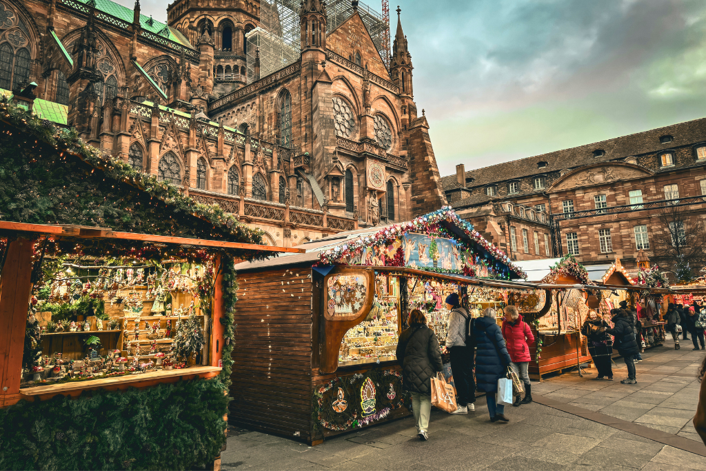 Kate browses festive stalls filled with ornaments and gifts at a European Christmas market set against the backdrop of a grand Gothic cathedral.