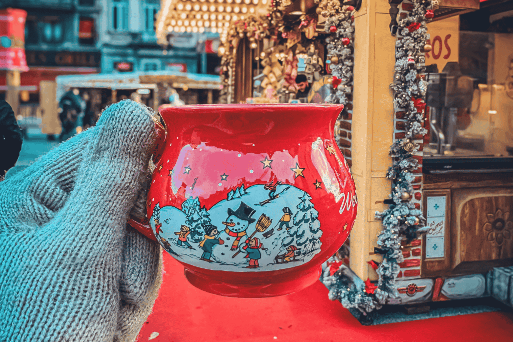 Close-up of a festive red mug with snowman and Christmas scene illustrations, held by a gloved hand in front of a decorated stall at the Brussels Christmas Market.


