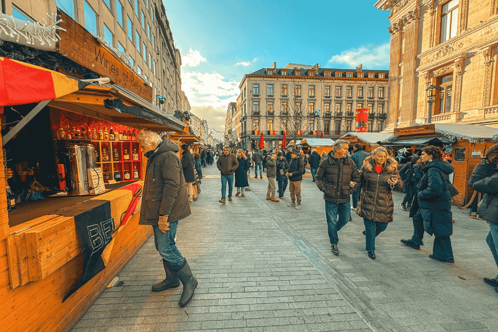 Crowds browse festive wooden stalls at Brussels Christmas Market, including a “Wines of Belgium” booth, with shoppers bundled up under a bright winter sky near ornate historic buildings.